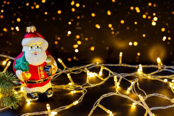 Toy Santa Claus among bright garlands on dark background with lights. New Year and Christmas background with copy space