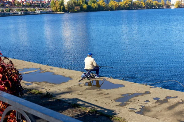 A man sits on a chair and fishes with a fishing rod on the city embankment. Sunny warm autumn day