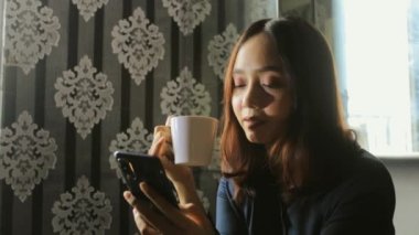 Rahatlamış Asyalı kadın iş için cep telefonu kullanırken kahve içiyor.