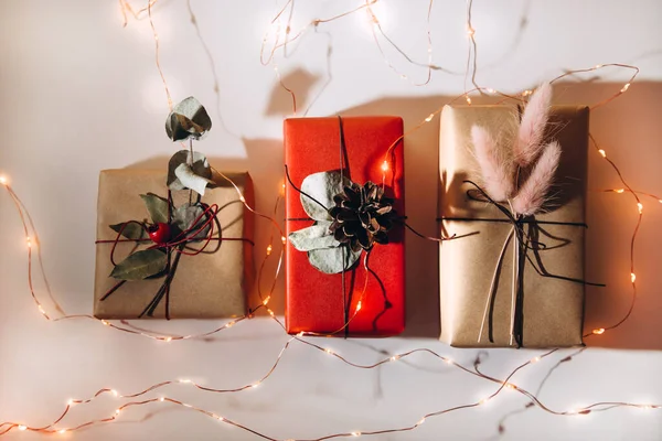 Piros és bézs ajándékok díszített száraz virágok és fenyőtoboz egy könnyű háttér karácsonyi fények — Stock Fotó