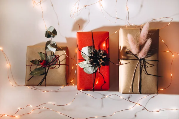 Presentes vermelhos e bege são decorados com flores secas e pinhas em um fundo leve com luzes de Natal — Fotografia de Stock