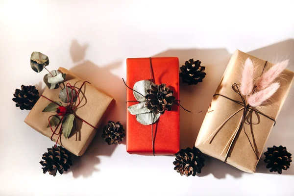 Les cadeaux sont décorés avec du pin chic et des fleurs séchées sur un fond blanc. Les cadeaux sont emballés dans du papier rouge et beige — Photo