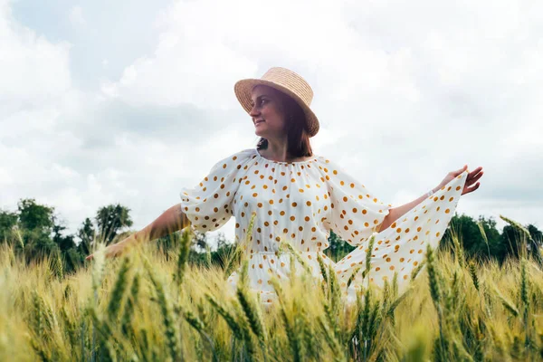 Das Mädchen im Weizenfeld trägt ein helles Kleid mit Tupfen und einen Strohhut — Stockfoto