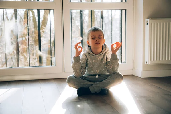 Çocuk yerde oturur ve pencerenin yanında meditasyon yapar. Güneş ışığıyla doludur.