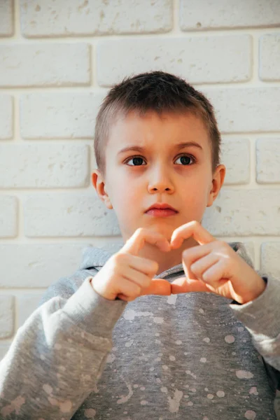 Chlapec ukazuje svým prstem srdce. Portrét dítěte proti zdi z bílých cihel — Stock fotografie