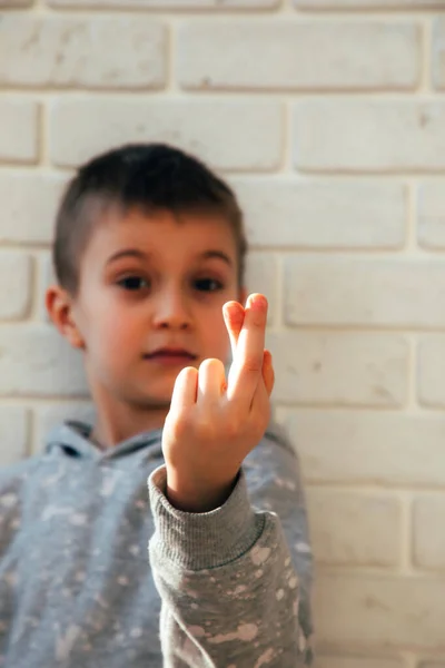O menino sorri e mostra um coração com os dedos. Retrato de uma criança contra uma parede de tijolo branco — Fotografia de Stock