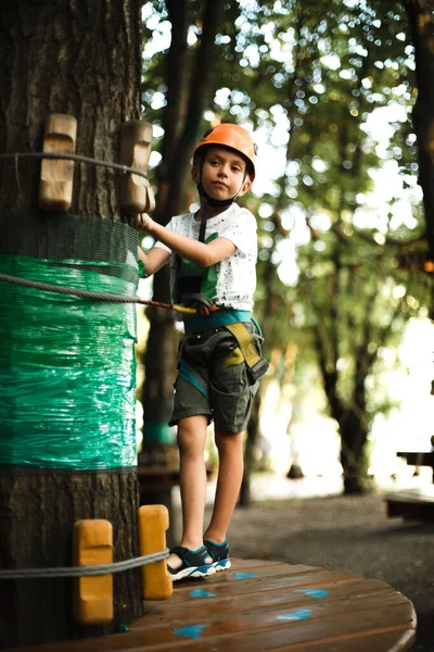 Un niño alegre en un casco naranja se divierte en el parque de cuerdas al atardecer — Foto de Stock