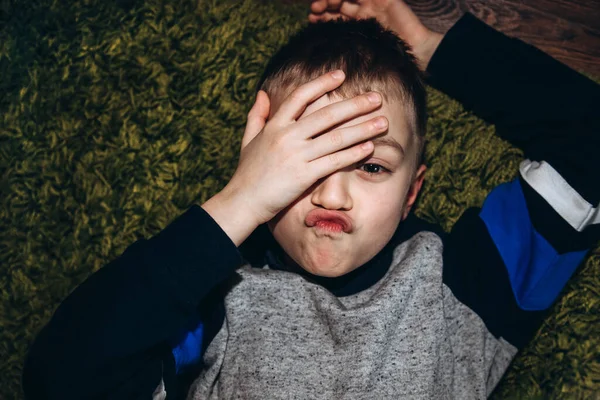 Portrait of a grimacing boy lying on the floor and covering his eye with one hand — Fotografia de Stock