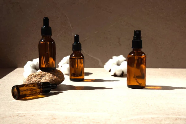 Clear amber bottle with natural cosmetics on a beige stone background with cotton flowers