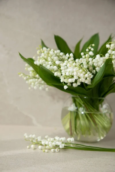 Bouquet of lilies of the valley in a round glass vase on a beige background. Front view