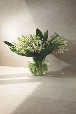 Bej arka planda yuvarlak cam bir vazoda vadideki zambaklar. Ön görünüm