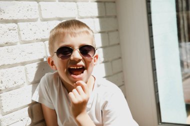 Güneş gözlüklü bir çocuk tuğla duvara yaslanmış oturuyor ve gülüyor. 6-7 yaşlarında bir çocuğun portresi.