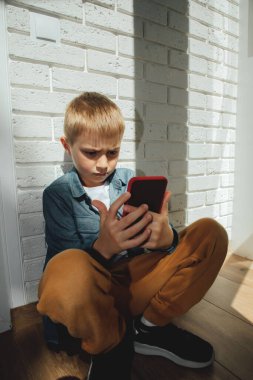 6-7 yaşlarında mavi gömlekli bir çocuk pencerenin yanında oturmuş akıllı telefona dikkatlice bakıyor..
