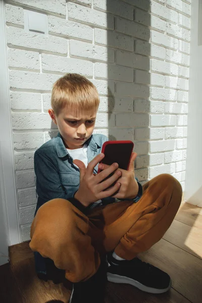 6-7 yaşlarında mavi gömlekli bir çocuk pencerenin yanında oturmuş akıllı telefona dikkatlice bakıyor..