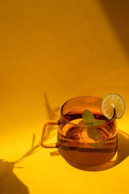 Sarı arka planda gölgesi olan bir dilim limonlu turuncu cam bardakta nane çayı. Ön görünüm