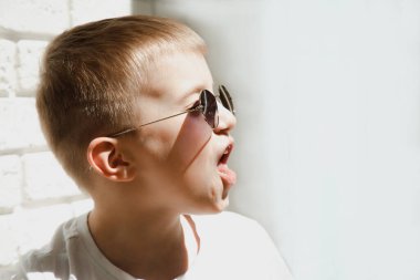 Güneş gözlüklü bir çocuk tuğladan duvara yaslanmış ve suratını buruşturup çığlık atıyor. 6-7 yaşlarında bir çocuğun portresi.