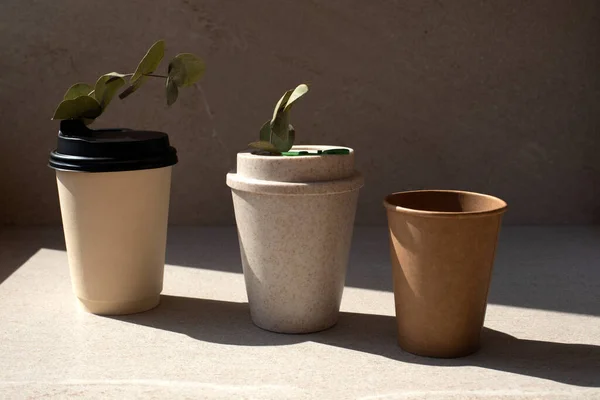 A set of cups made of eco-materials with eucalyptus branches. Disposable and reusable eco cups on a stone beige background. Front view