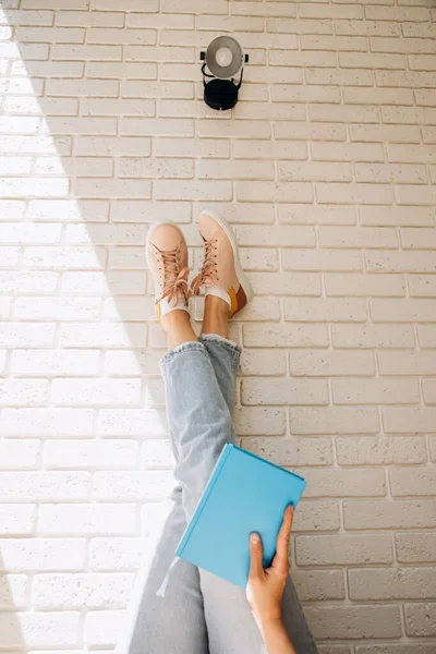 Woman holding an close book with her legs up in blue jeans and pink sneakers against a brick wall background