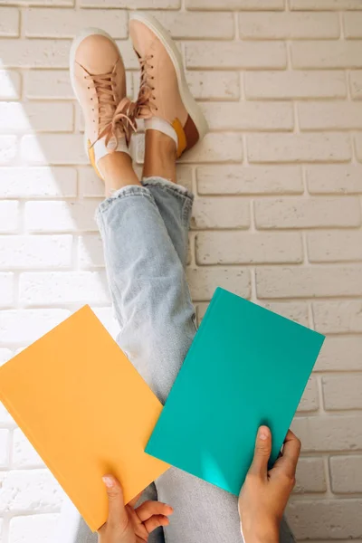 Woman holding an two close books or notebooks with her legs up in blue jeans and pink sneakers against a brick wall background