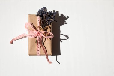 A gift box in kraft paper tied with a pink lace ribbon and lavender flowers on a white background with a shadow. Top view