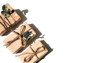 Kraft kağıdında el yapımı hediyeler, geniş bir iple bağlanmış, beyaz bir masada kurumuş çiçeklerle süslenmiş. Üst görünüm