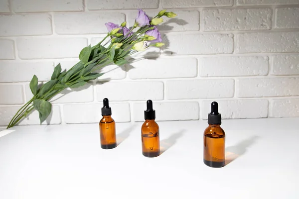 A group of amber bottle on a white table with face serum products near purple eustoma. Glass bottle with self care product. Front view