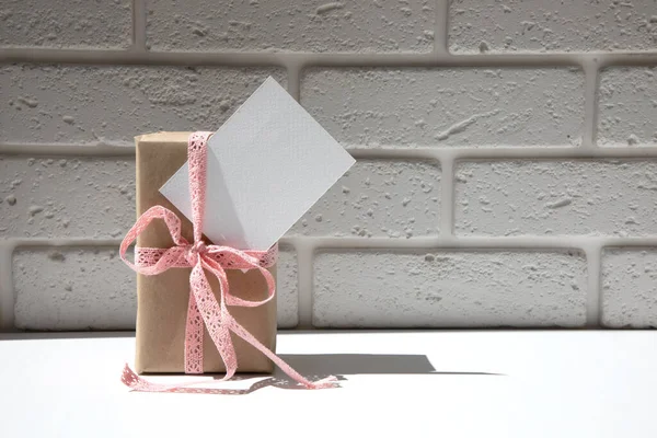 A gift box in kraft paper tied with a pink lace ribbon and a white postcard on a white background with a shadow. Front view