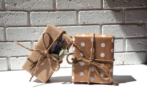 Два подарка ручной работы из крафтовой бумаги, обвязанные широким шпагатом, украшенные сухими цветами на белом столе. Вид спереди