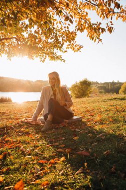 Beautiful girl with long blond hair in autumn landscape at sunset. Autumn portrait of a girl sitting on the grass, front view, selective focus