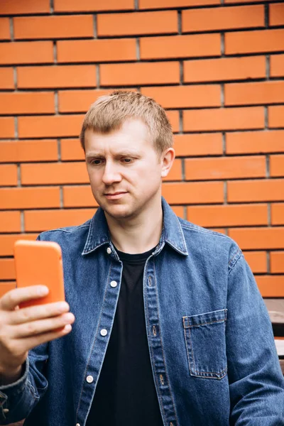 A man in a casual jacket sits on a bench and takes a selfie. Portrait of a boy against a background of a red brick wall, front view