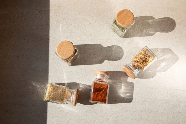A group of seasoning in glass jars on a light stone background with shadows. Paprika, herbs, mustard, garlic, flat lay