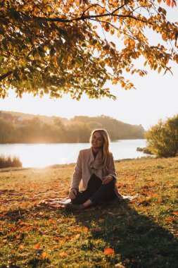 Beautiful girl with long blond hair in autumn landscape at sunset. Autumn portrait of a girl sitting on the grass, front view, selective focus