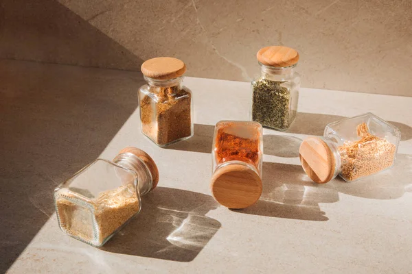 A group of seasoning in glass jars on a light stone background with shadows. Paprika, herbs, mustard, garlic, front view, selective focus