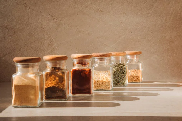 A group of seasoning in glass jars on a light stone background with shadows. Paprika, herbs, mustard, garlic, front view, selective focus