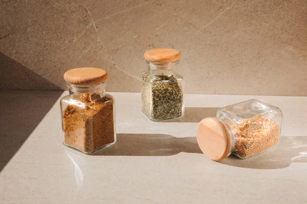 A group of seasoning in glass jars on a light stone background with shadows. Paprika, herbs, mustard, garlic, front view, selective focus