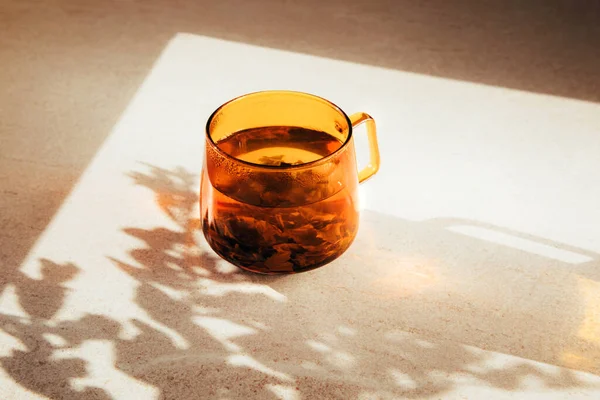 Glass transparent orange mug with hot green tea, shadow and reflection on light stone background. Front view