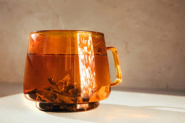 Glass transparent orange mug with hot green tea, shadow and reflection on light stone background. Front view