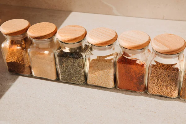 A group of seasoning in glass jars on a light stone background with shadows. Paprika, herbs, mustard, garlic, front view, selective focus