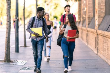 Farklı etnik kökenlere sahip öğrenciler okul malzemeleriyle sokakta yürüyorlar.