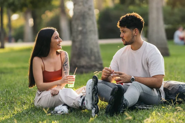 Coppia seduta sull'erba di un parco che beve soft drink indossando pattini in linea — Foto Stock