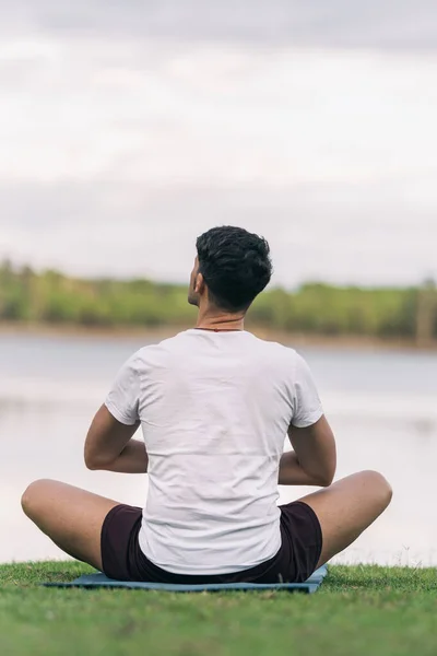 Achterzijde van een atletische man zittend op een yoga mat tegenover een meer — Stockfoto