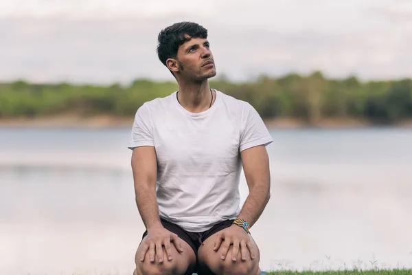 Yoga minderinin üzerinde oturan atletik bir adam, dikkati dağılmış bir ifadeyle gölün yanında.