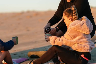 Adam yoga dersinden sonra kumsalda oturan bir kadına çay dolduruyor.