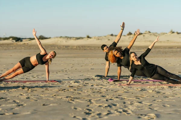 Тренер по йоге и группа мультиэтнических учеников, занимающихся боковой доской на пляже — стоковое фото