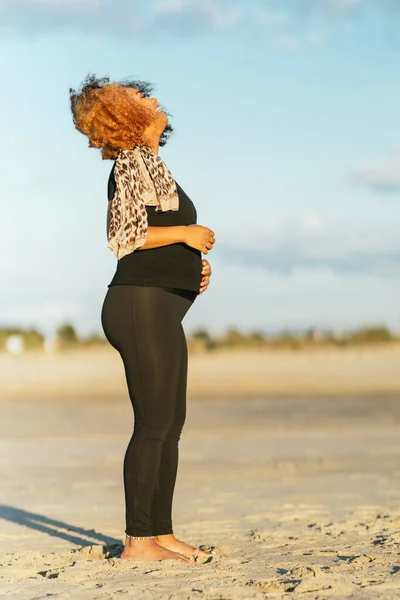 Profil över en gravid latinamerikansk kvinna som står på stranden i sportkläder — Stockfoto