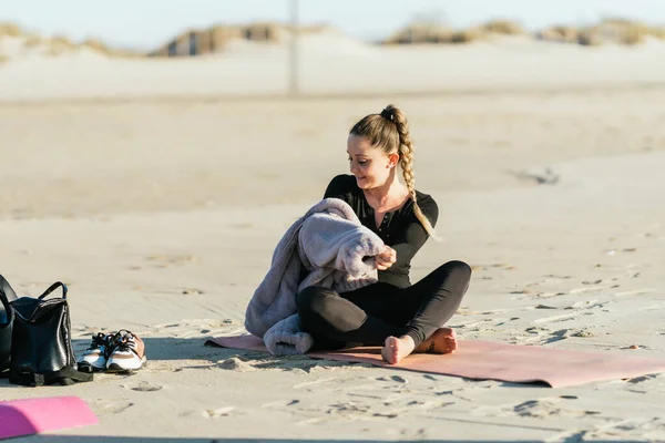 Женщина снимает свитер, сидя на коврике для йоги на пляже — стоковое фото