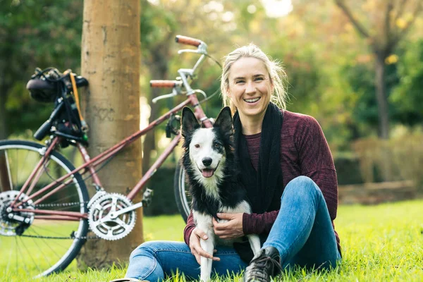 Mulher e um cão sentado na grama de um parque ao lado de uma bicicleta — Fotografia de Stock