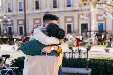 Two multiethnic friends hugging while holding bags with presents outdoors