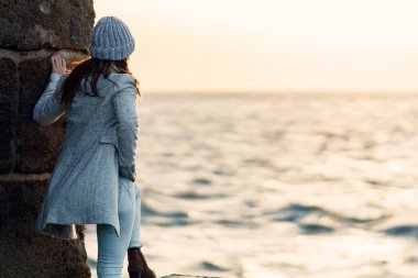 Yünlü şapkalı kadın, güneşin denize batışını izlerken ayağını duvara dayar.