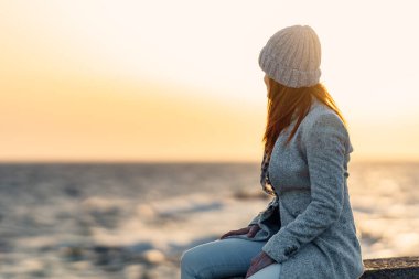 dalgakıranda oturmuş okyanus ufkuna bakan kızıl saçlı bir kadının arka ışıklandırması.